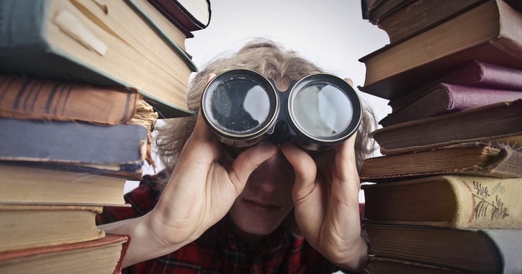 Anonymous person with binoculars looking through stacked books