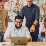 Design review in startup office with macbook and artwork. Man wearing Apple Watch. Joyful team working together and laughing.