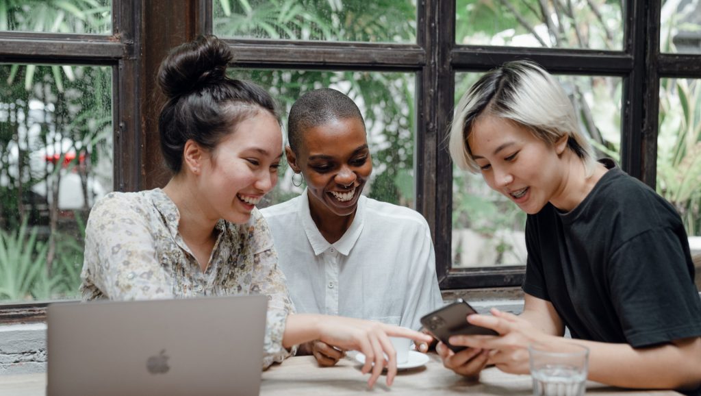 Happy multiethnic women having fun while using gadgets in cafeteria