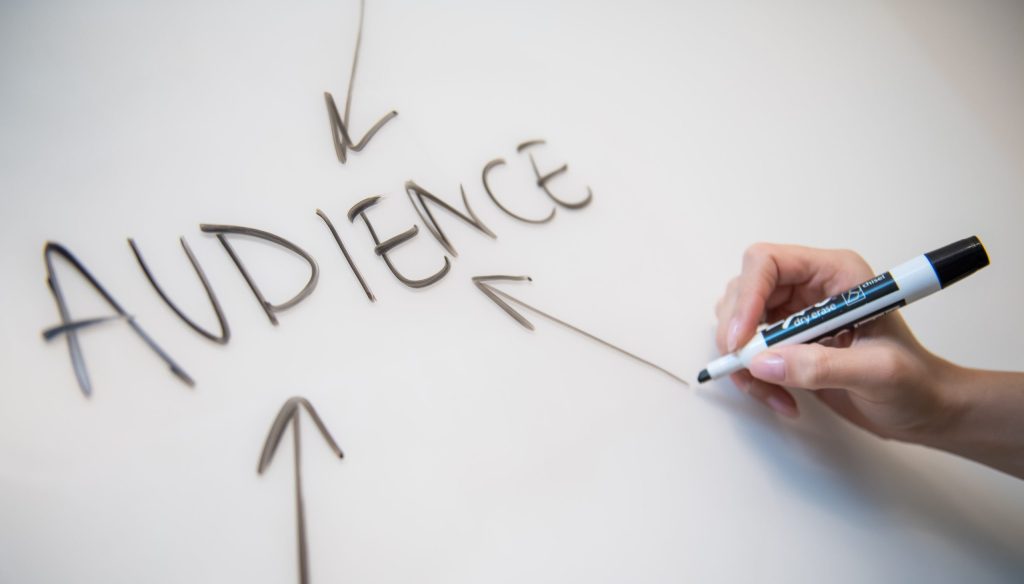 Woman's hand writing the word "audience" on a whiteboard, with arrows.